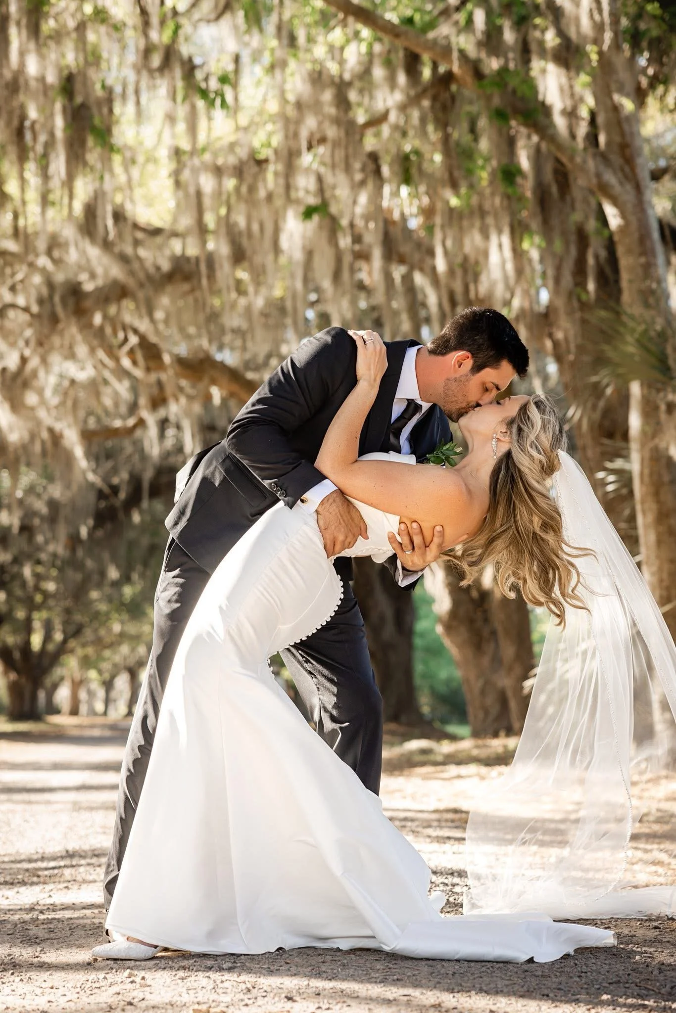A bride and groom sharing a kiss outdoors during their wedding. The groom is in a black suit, and the bride is in a white wedding dress with long hair. The groom is holding the bride in a dip, and they are kissing under a tree with hanging moss.