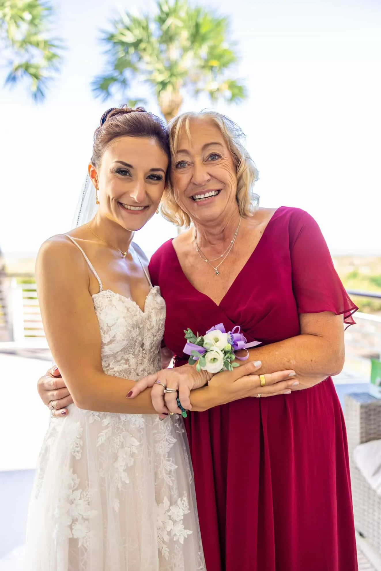A bride in a lace wedding dress and an older woman in a red dress are smiling and embracing outdoors, with a palm tree in the background, during a wedding celebration.