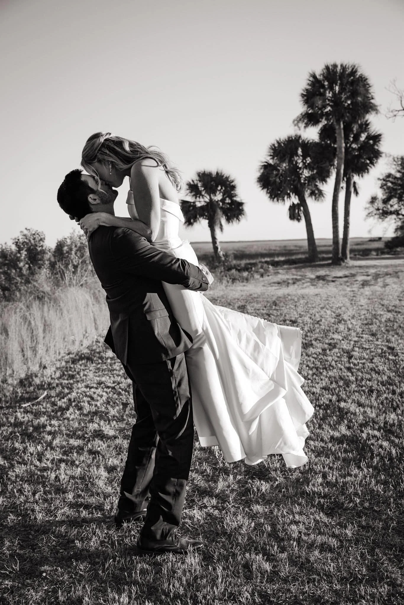 A man in dark clothing carrying a woman in a white dress during sunset in a grassy field with palm trees in the background.
