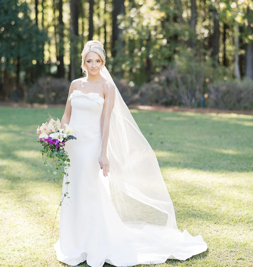 A bride in a white wedding gown holding a bouquet of flowers, standing outdoors on a grassy area with trees in the background.