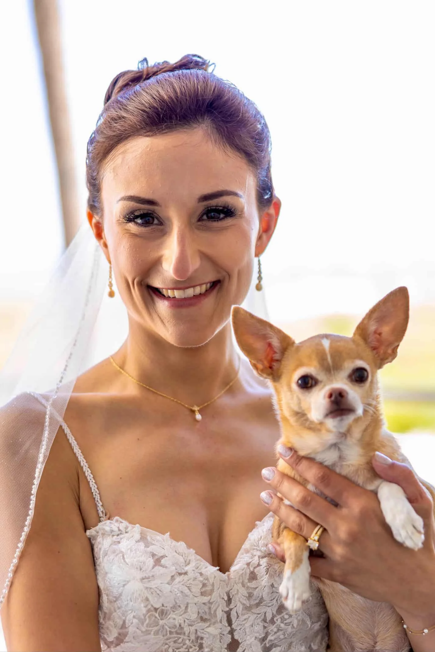 Close-up of a smiling woman in wedding attire holding a small tan and white dog.