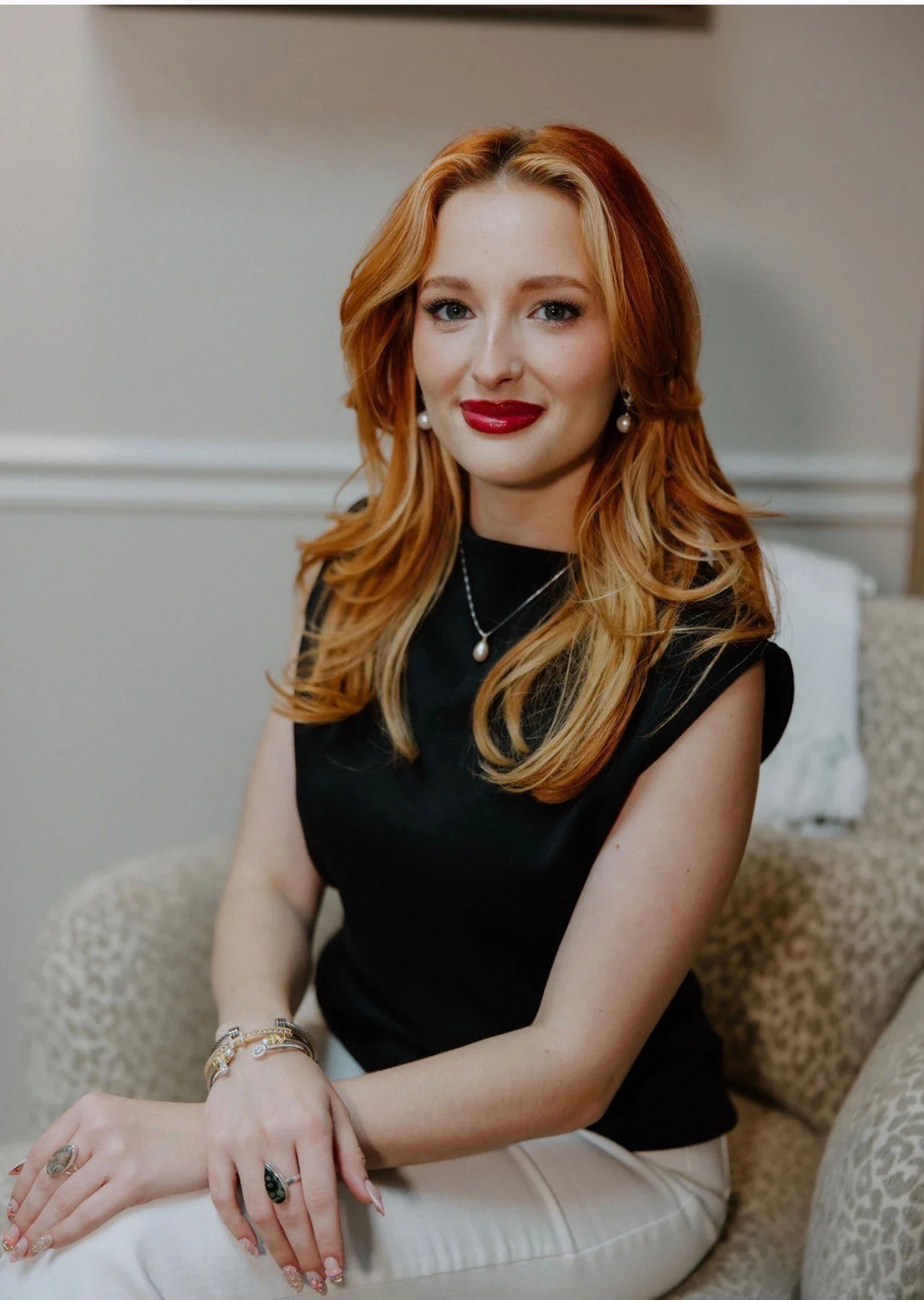 A woman with long, wavy red hair, wearing a black sleeveless top, pearl earrings, and a pearl necklace, sitting on a patterned armchair in a room with beige walls.