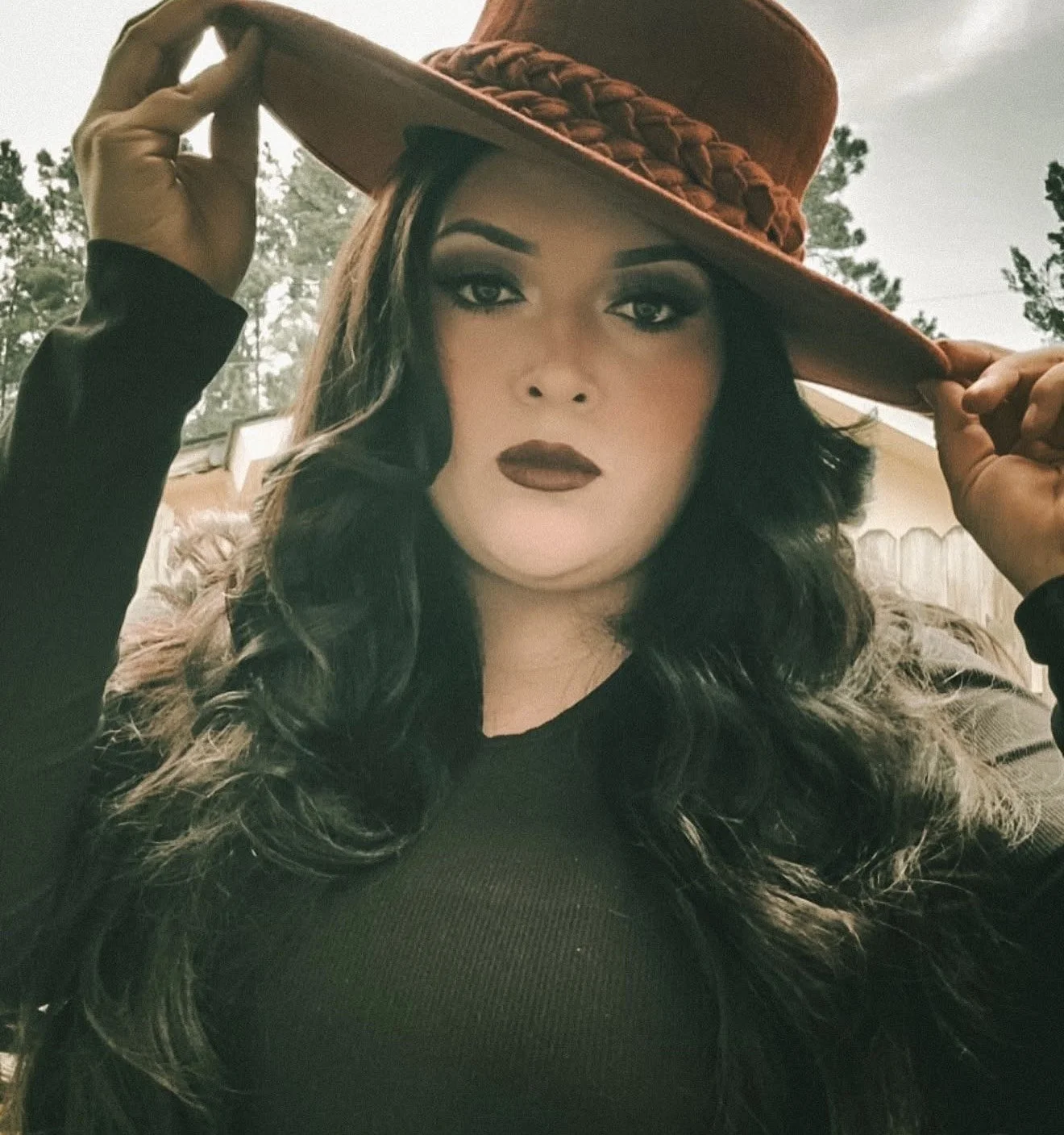 Woman with long wavy black hair wearing dark makeup and a black shirt, holding a brown wide-brimmed hat with a braided band outdoors.