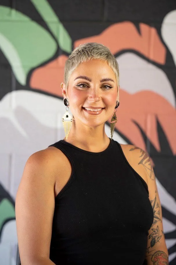 A woman with short blonde hair and tattooed arm, smiling, wearing a black sleeveless top and large earrings, standing in front of colorful abstract wall art.