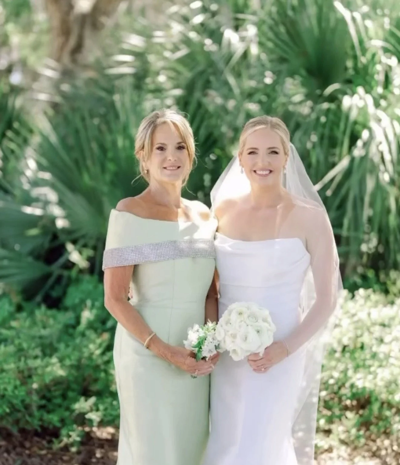 Two women, one in a green dress and the other in a white wedding dress, smiling and standing outdoors among green plants, holding bouquets of white flowers.