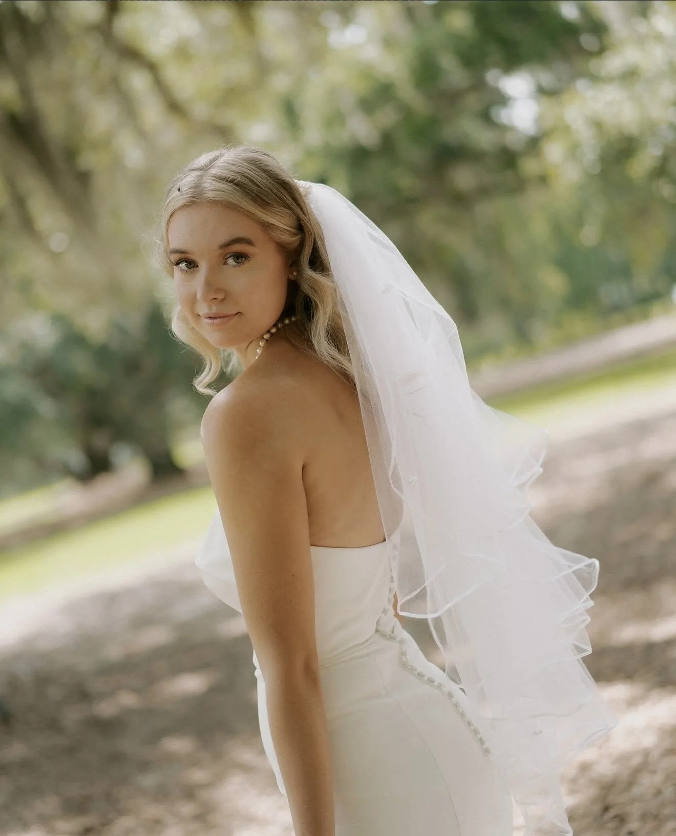A bride with blonde hair and a pearl necklace, wearing a white strapless wedding gown and veil, standing outdoors with blurred green trees in the background.