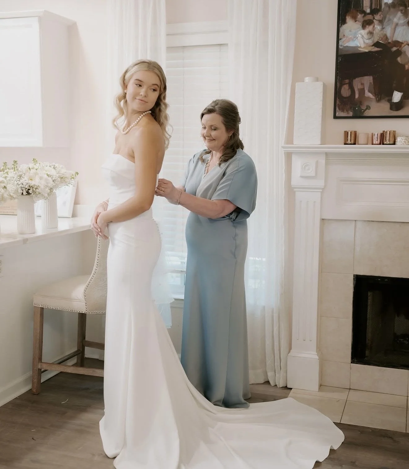 A woman in a wedding dress getting help from another woman in a light blue dress, likely during a wedding preparation in a cozy, well-lit room with white walls and decor.