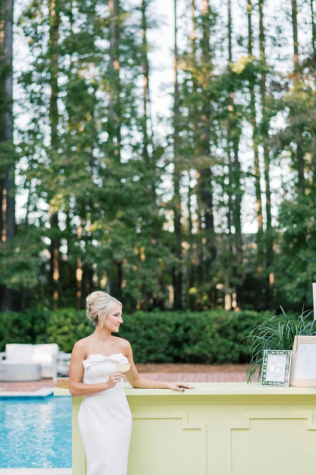 A woman in a white strapless dress holding a cocktail near a pool, standing at a light green bar with framed photos and menu, with a wooded background.