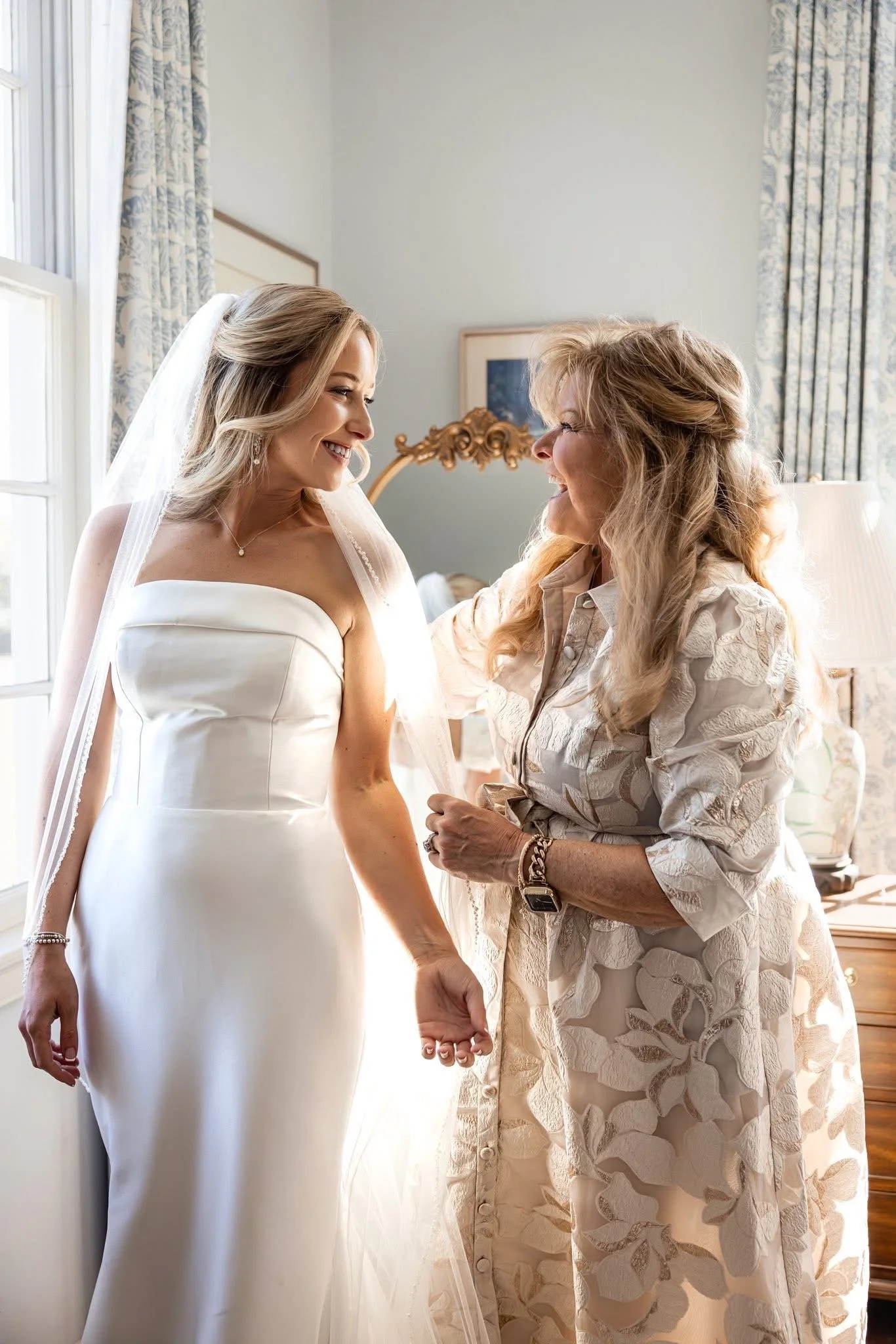 A bride in a white wedding dress and veil holding hands with an older woman in a cream-colored dress with floral embroidery, both smiling and looking at each other in a sunlit room.