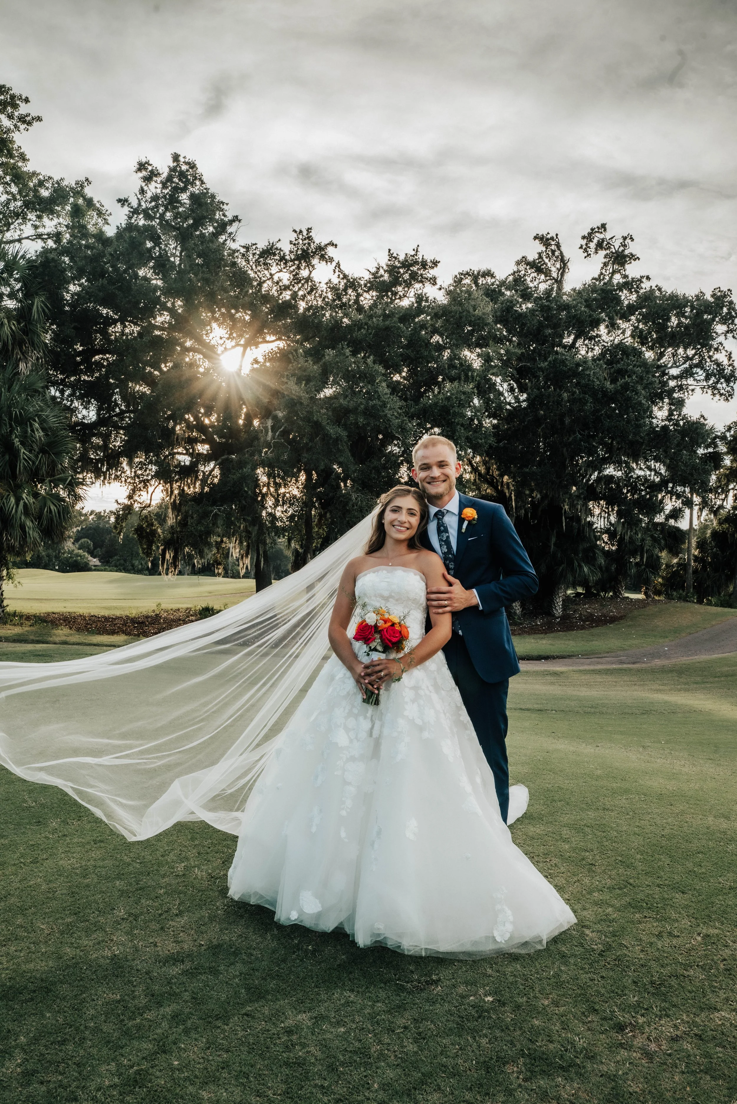 A bride and groom smiling and posing together outdoors on a cloudy day, with the bride wearing a white wedding gown and veil, holding a bouquet of red, orange, and yellow flowers, while the groom is dressed in a blue suit with a boutonniere. They are