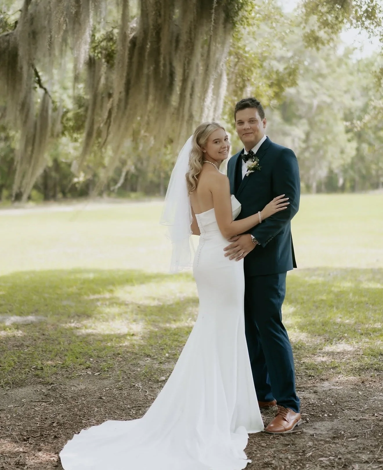 A bride and groom standing outdoors on a grassy area with large trees in the background, dressed in wedding attire, smiling and embracing.
