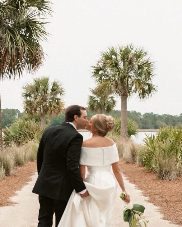 A newly married couple walking down a sandy path lined with palm trees, with the bride holding a bouquet of flowers, during daytime with an overcast sky.