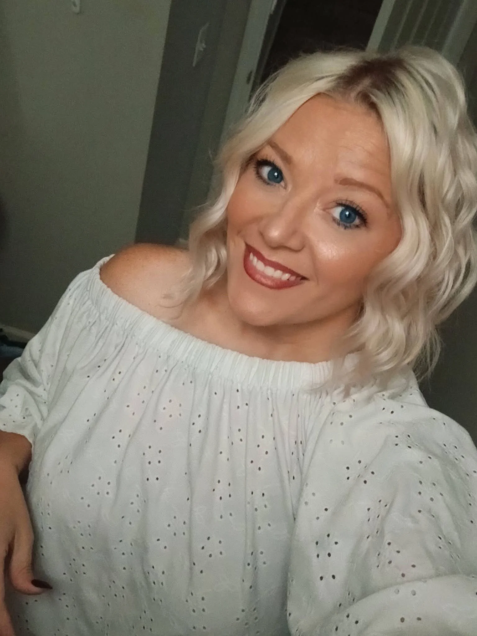 A woman with curly blonde hair and blue eyes smiling, wearing a white off-shoulder top with small black polka dots.