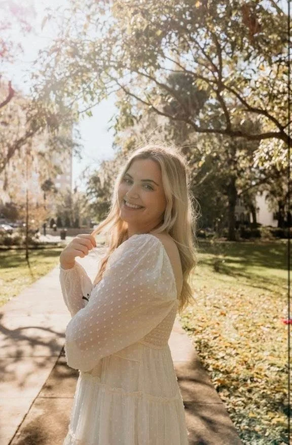A young woman with long blonde hair smiling and looking at the camera, standing on a sidewalk in a park with trees and fallen leaves, during autumn.