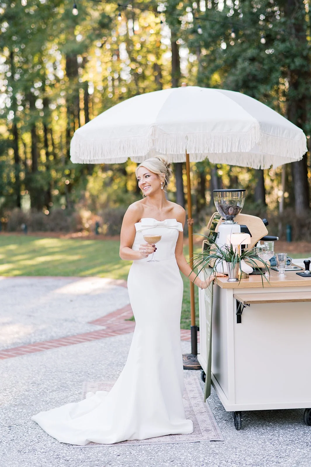 A woman in a white wedding dress holding a cocktail stands under a large white parasol outdoors, with a coffee or beverage station to her right, surrounded by trees and greenery.