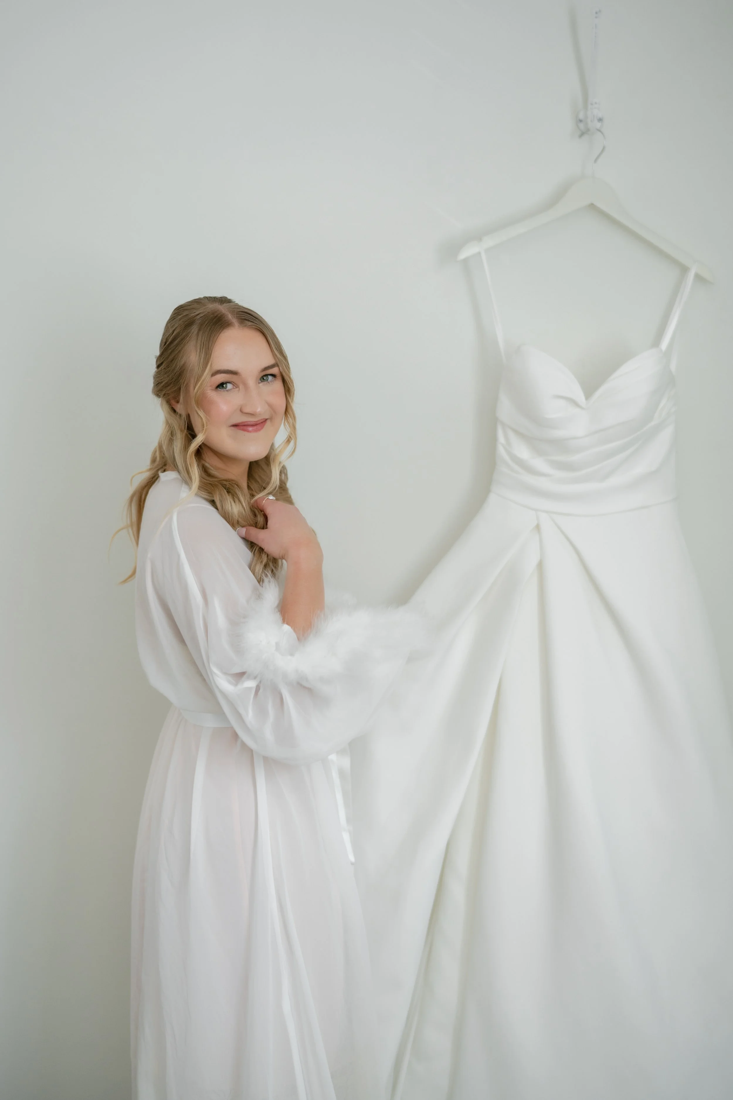 A young woman with long wavy blonde hair, dressed in a white gown with fluffy sleeves, smiling and holding her hand near her shoulder. Behind her is a white bridal dress on a hanger hanging from the ceiling.