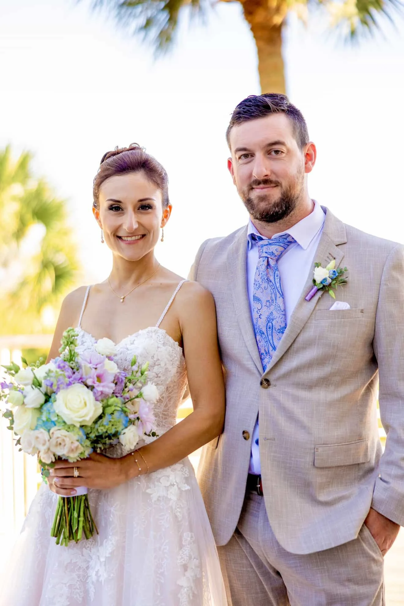 A smiling bride in a white lace wedding dress holding a bouquet of white, pink, and purple flowers, standing next to a groom in a beige suit with a light blue dress shirt and a patterned blue tie, outdoors with palm trees in the background.
