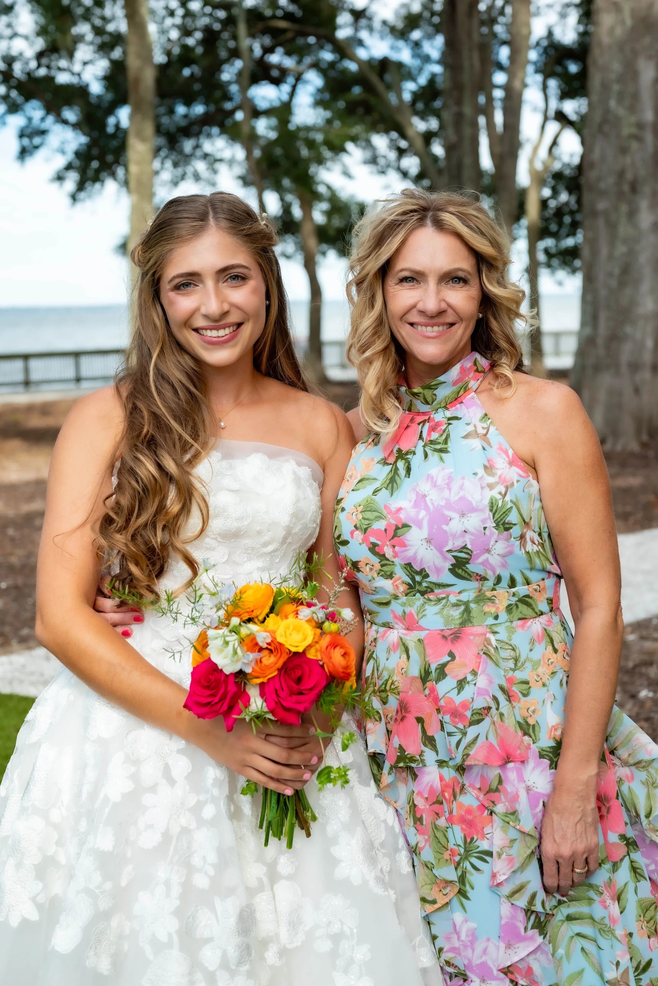 A young woman in a wedding dress holding a bouquet of colorful flowers stands next to an older woman in a floral dress outdoors surrounded by trees.