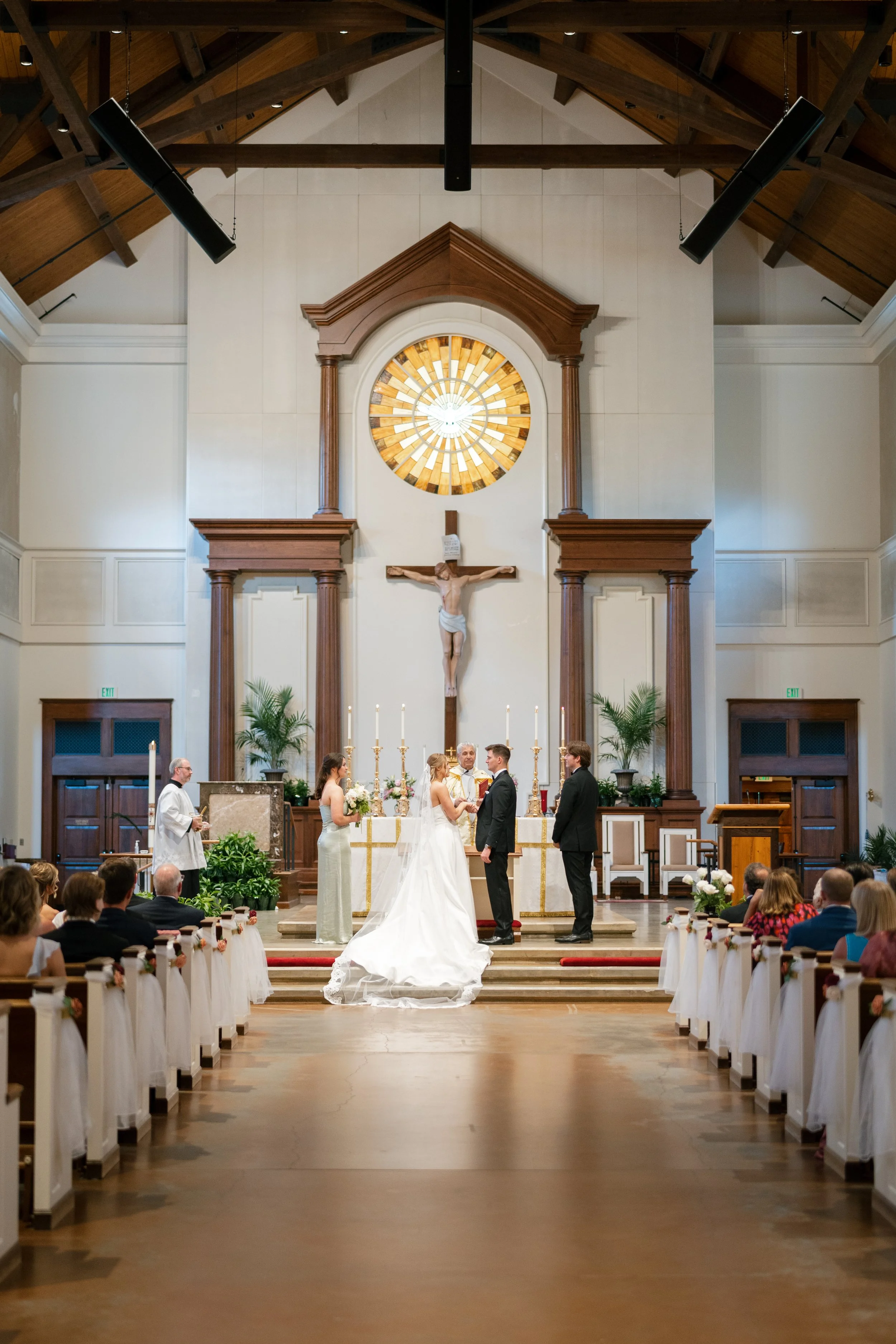 A wedding ceremony inside a church with a clergy officiant and a bride and groom standing before him. Guests seated on either side, church features stained glass window with a circular design behind the altar, wooden trusses on ceiling, and floral ar
