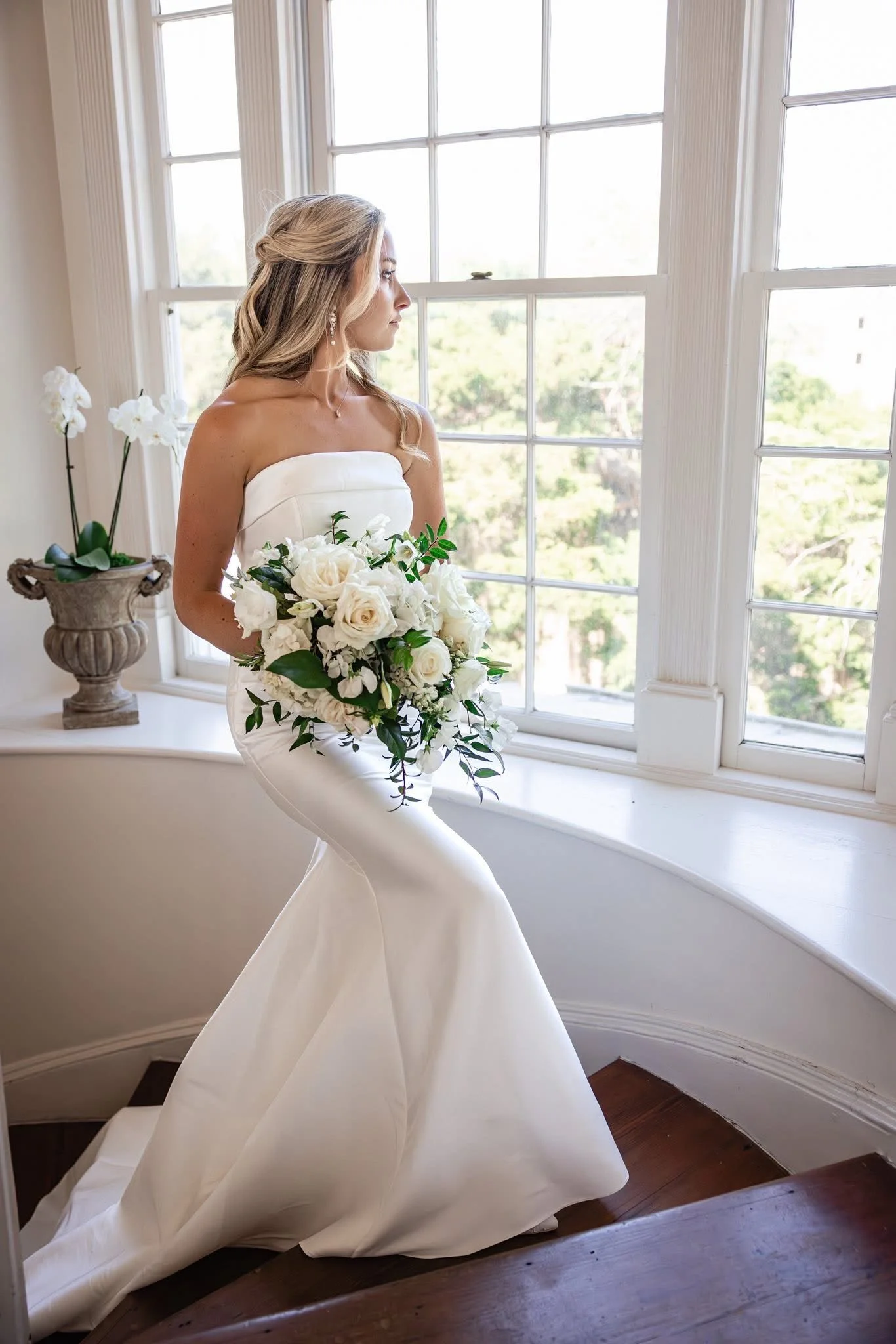A woman in a wedding dress holding a bouquet of white roses and greenery, sitting by a large window with white frames and a view of trees outside.