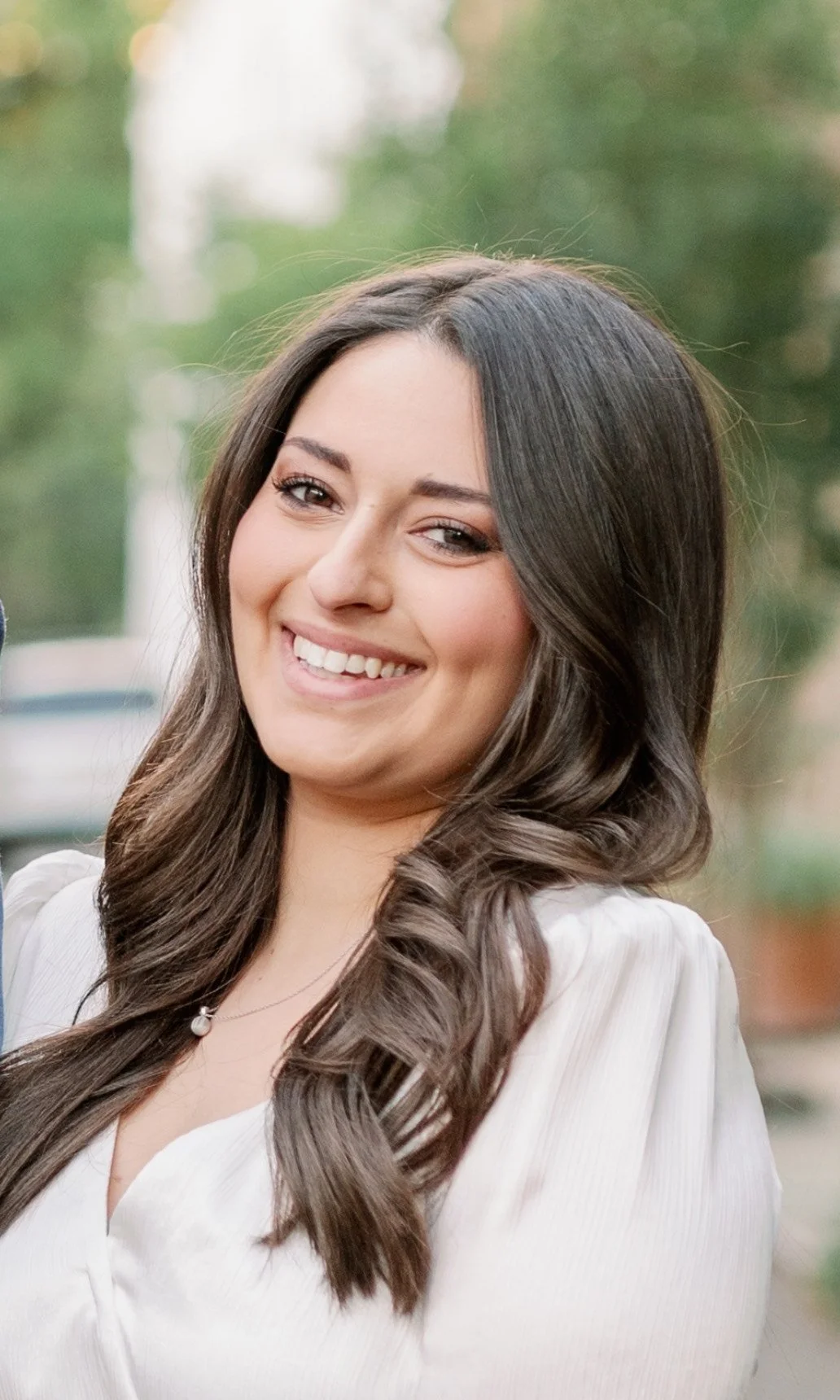 A smiling young woman with long, dark, wavy hair outdoors, wearing a white top and a necklace with a pearl pendant.