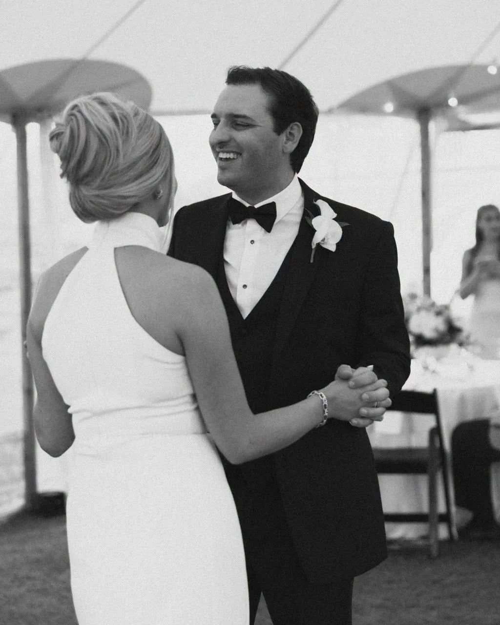 A smiling groom in a tuxedo with a boutonniere dancing with a woman in a white dress at a wedding reception under a white tent.