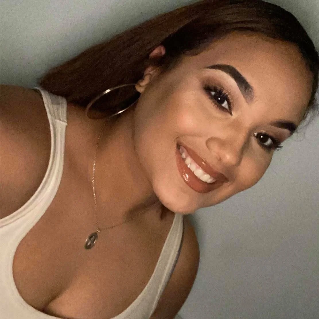 A young woman smiling with long brown hair, wearing a white tank top, hoop earrings, and a necklace, posing against a plain background.
