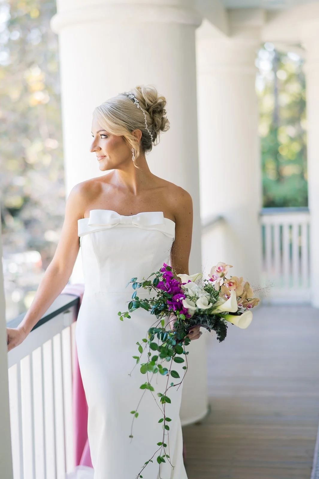 A bride in a white strapless wedding dress holding a bouquet of purple, white, and pink flowers, standing on a porch with white columns and a white railing, looking to the side.