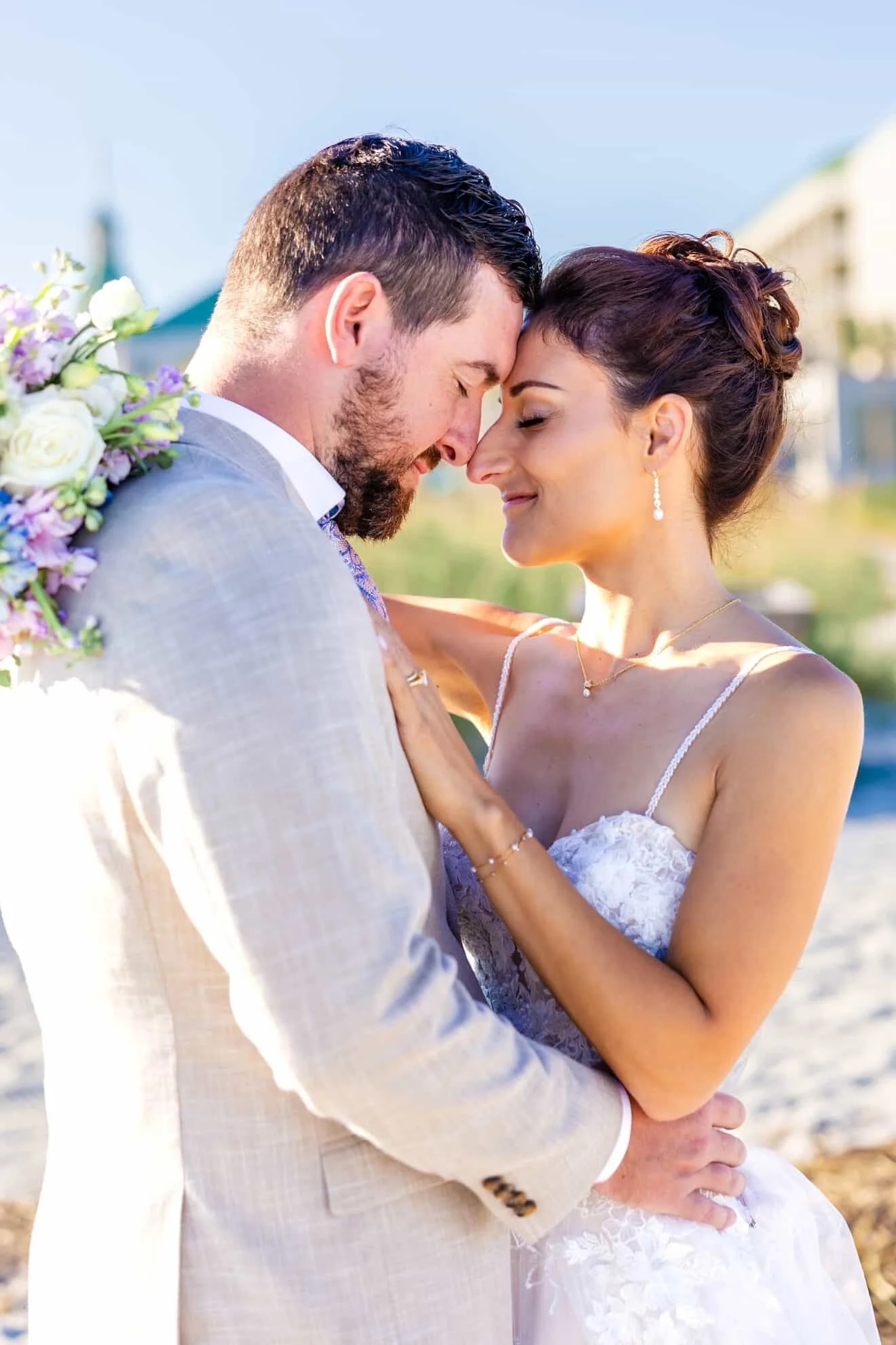 A bride and groom holding each other with their foreheads touching outdoors on a sunny day, with the groom carrying a bouquet of flowers.
