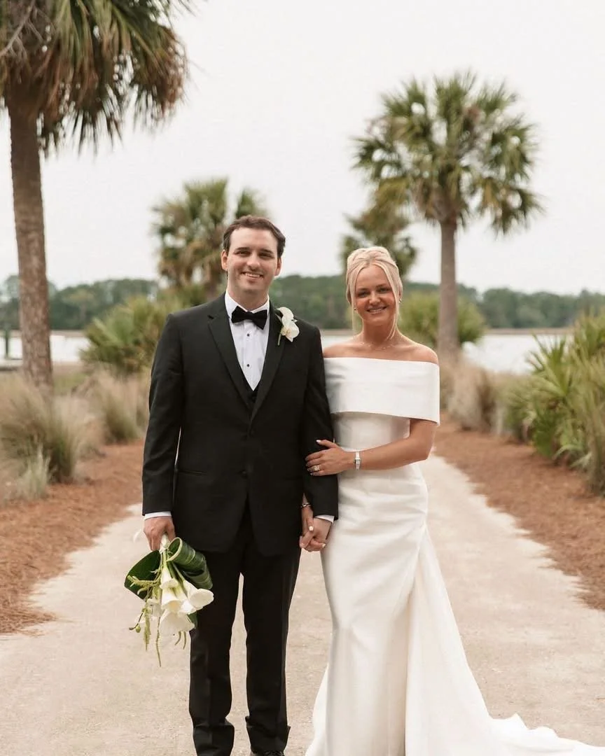 A newlywed couple standing on a path lined with palm trees by a body of water, outdoors during the daytime, smiling at the camera.