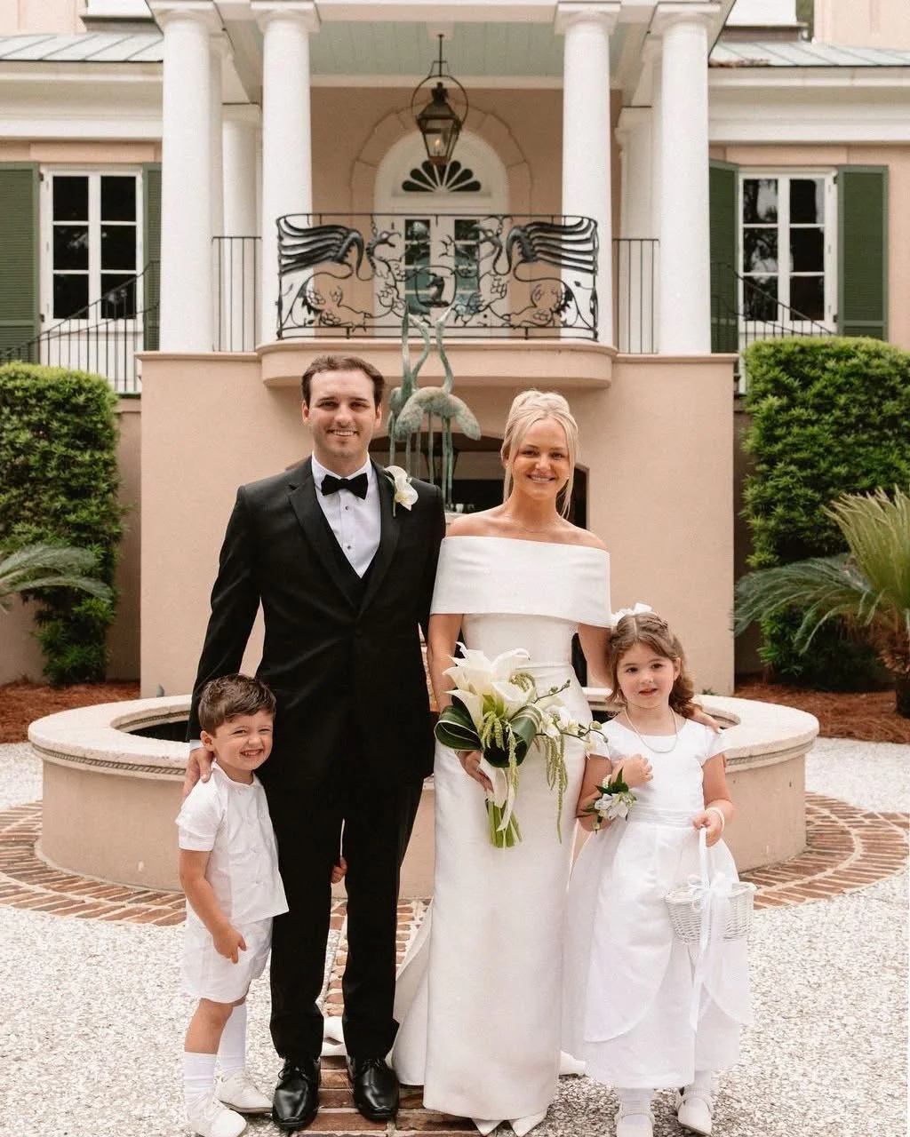 A wedding party with a groom in a black tuxedo, a bride in a white wedding dress holding a bouquet, a young boy in white shorts and shirt, and a young girl in a white dress with a basket, standing outdoors near a beige fountain with a house and green