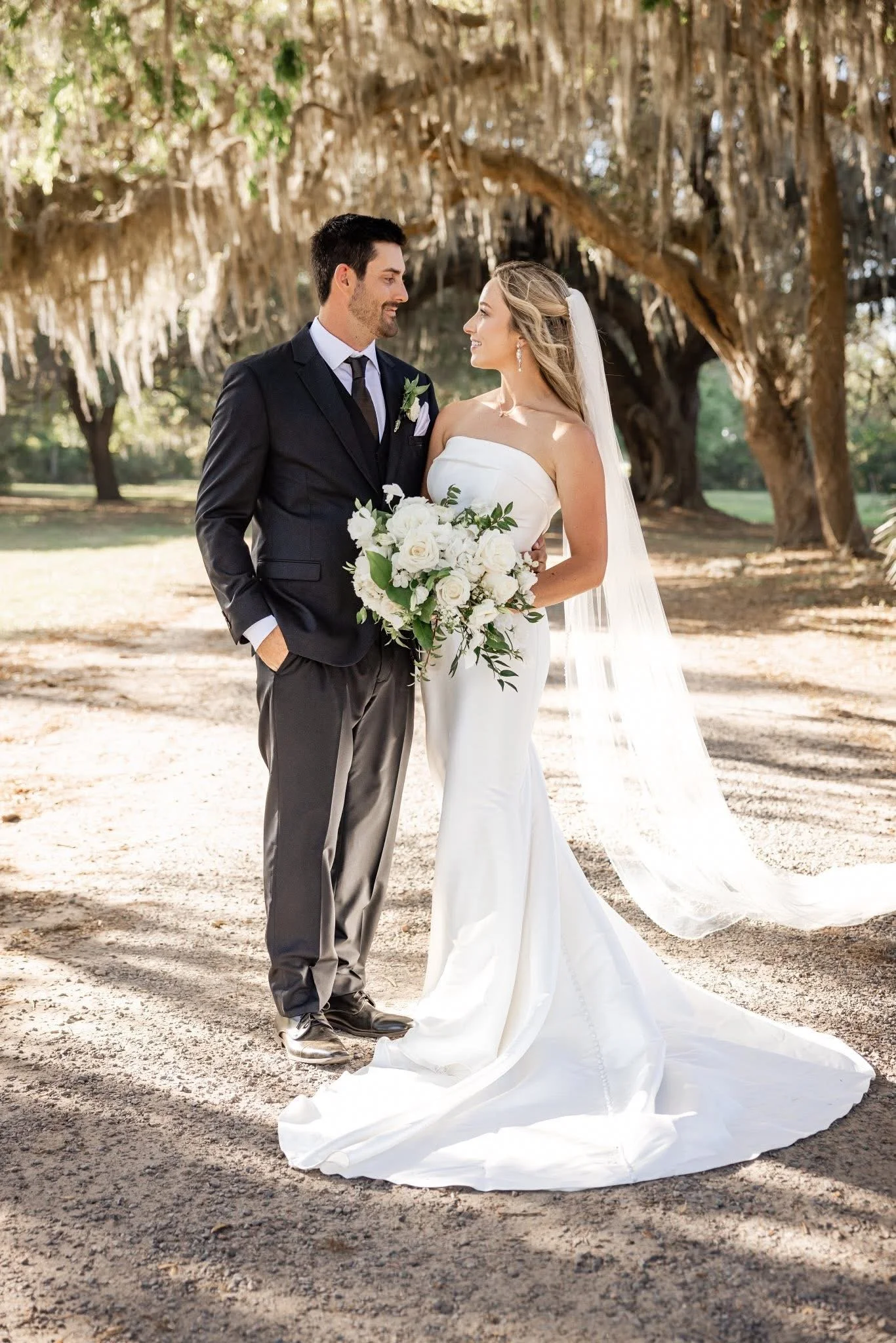 A bride and groom standing close together outdoors under a large, leafy tree on a sunny day. The groom is dressed in a dark suit and tie, smiling at the bride. The bride is wearing a strapless white wedding gown with a long train and veil, holding a 