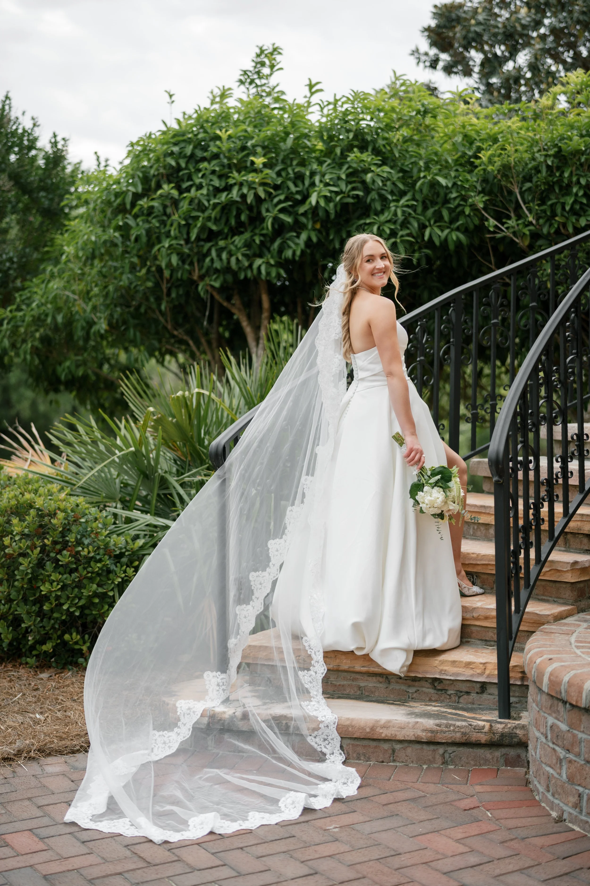 A bride in a wedding dress stands on outdoor stairs with a black iron railing, holding a bouquet of white flowers, with a large green leafy bush and trees in the background.