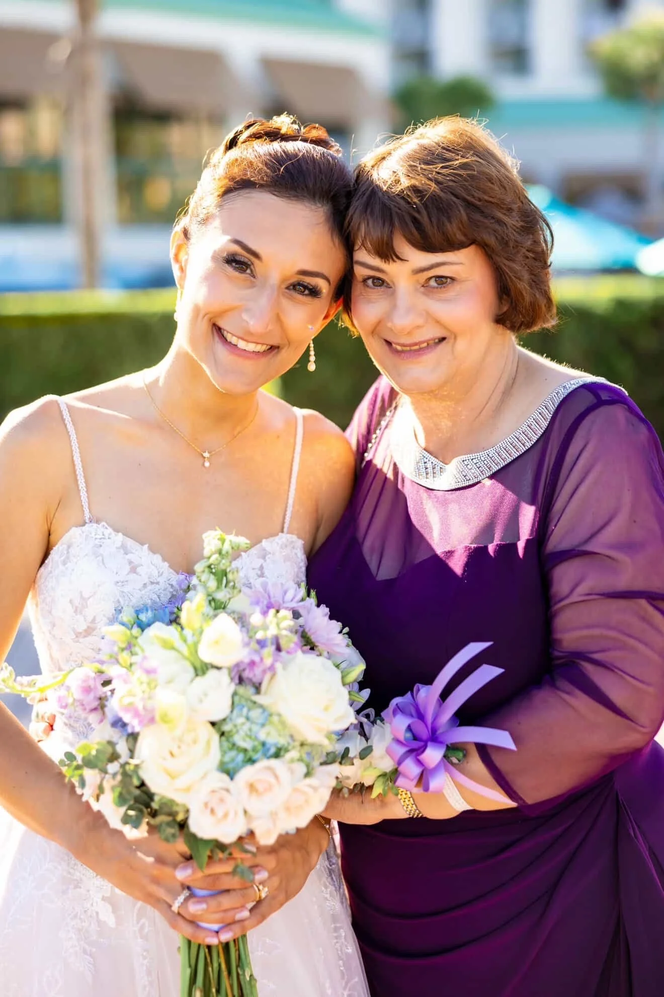 A bride holding a bouquet of flowers standing next to an older woman in a purple dress, both smiling and with their heads close together outdoors.