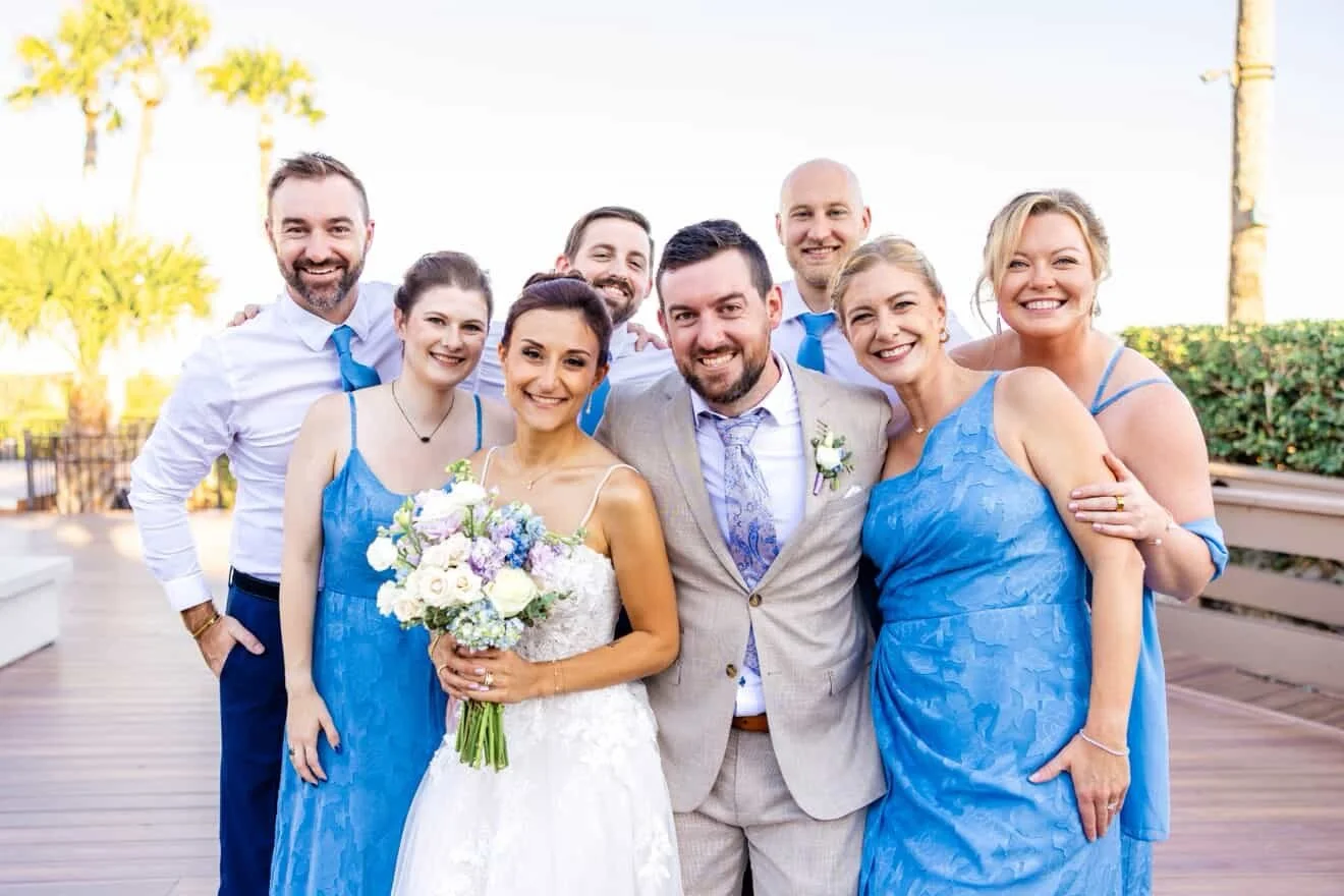 Group of nine people, nine smiling, including a bride in a white wedding dress holding a bouquet, and an officiant in a light-colored suit, outdoors on a sunny day.