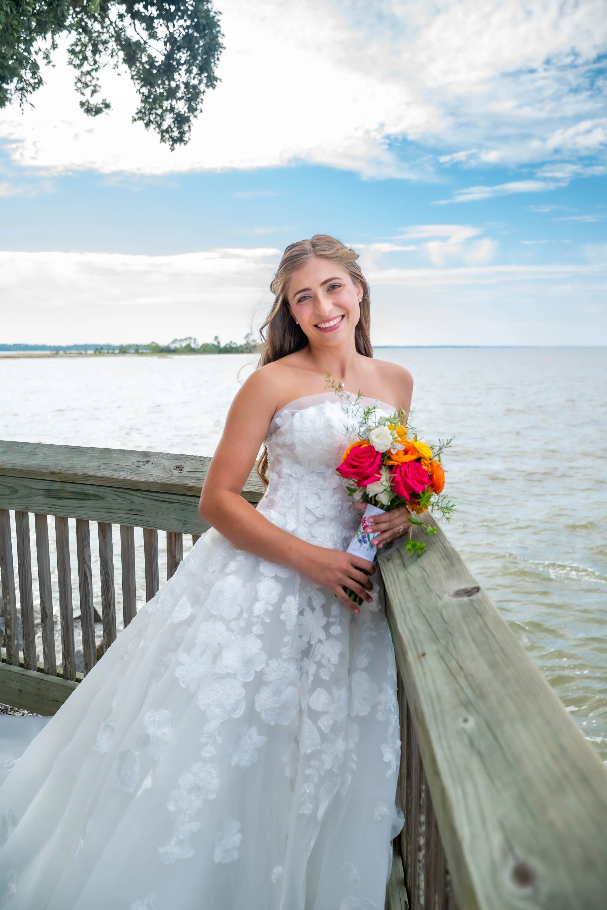 A smiling woman in a white wedding dress holding a colorful bouquet standing on a wooden pier by the water, with trees and a partly cloudy sky in the background.