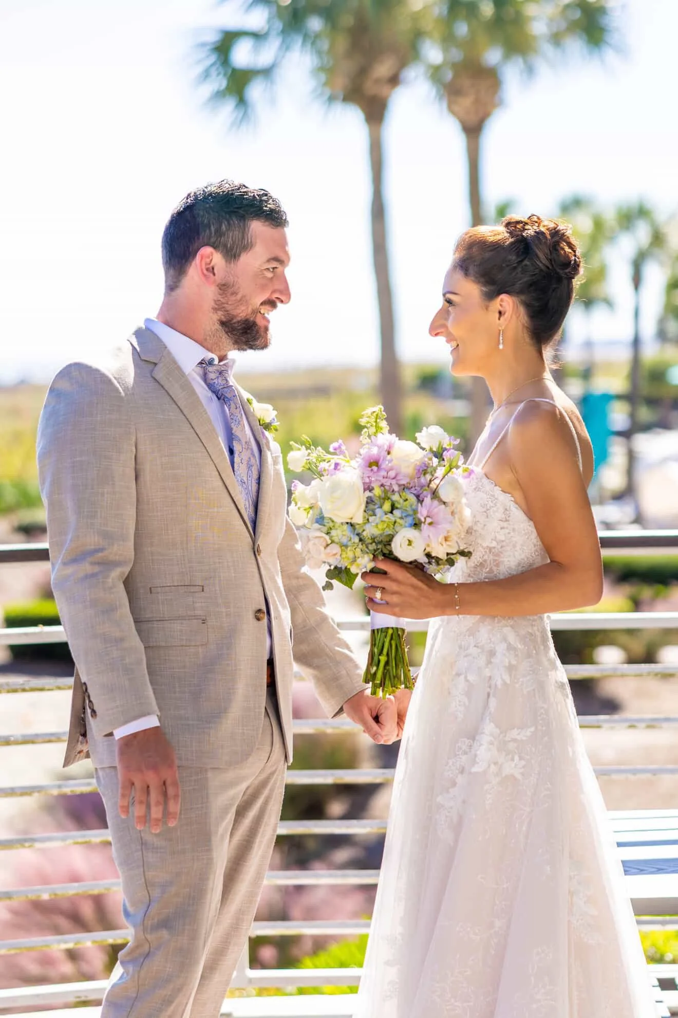 A bride and groom stand face to face outdoors, holding hands, with the bride holding a bouquet of white and pink flowers. The groom wears a light-colored suit, and the bride wears a white lace wedding dress with an updo hairstyle. There are palm tree