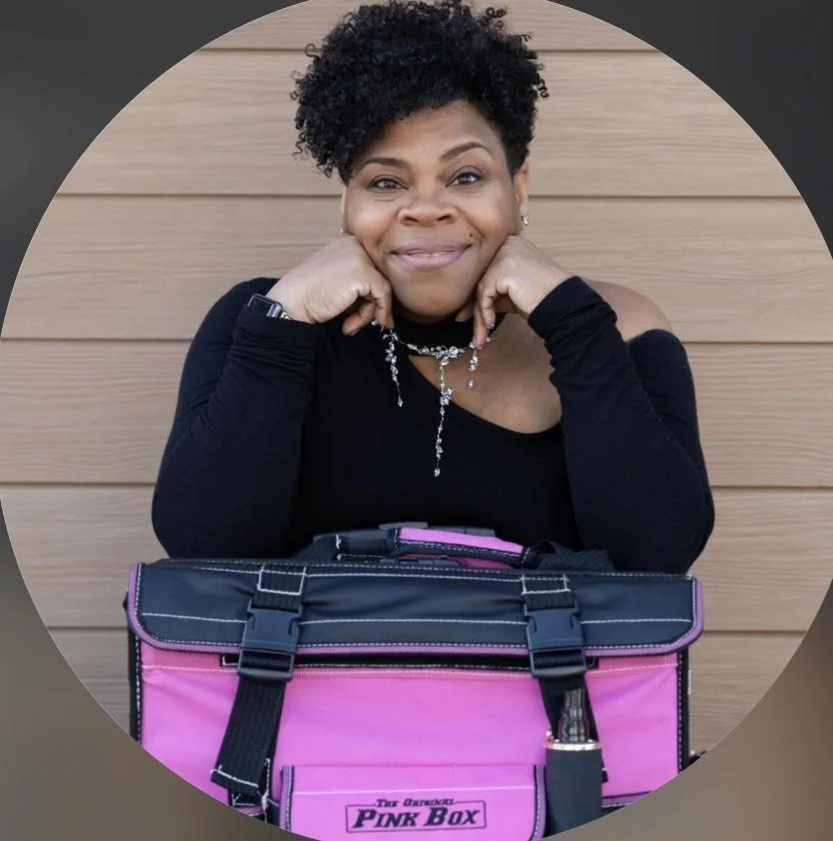 Smiling woman with short curly hair posing behind a pink and black lunchbox labeled 'Pink Box' in front of a wooden wall.