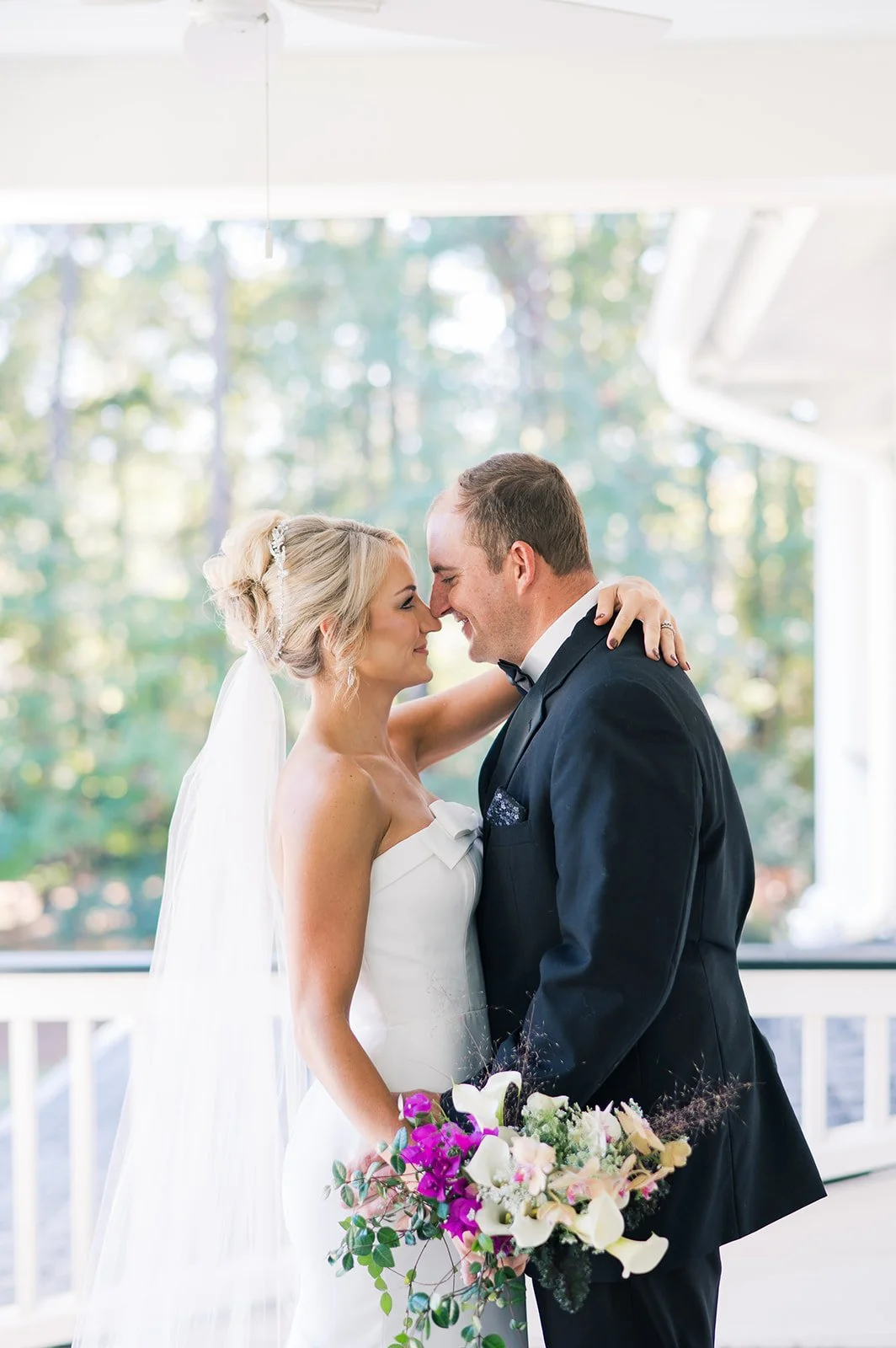 A bride and groom are smiling and touching foreheads during their wedding, with a bouquet of flowers in the bride's hand and a bright, outdoor background.