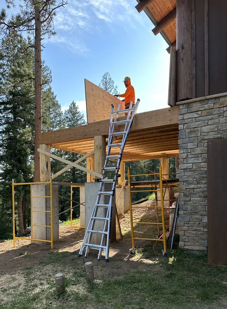 A construction worker in an orange shirt and cap working on a wooden deck or roof extension of a house, with a ladder leaning against the structure, surrounded by scaffolding, in a wooded area on a sunny day.