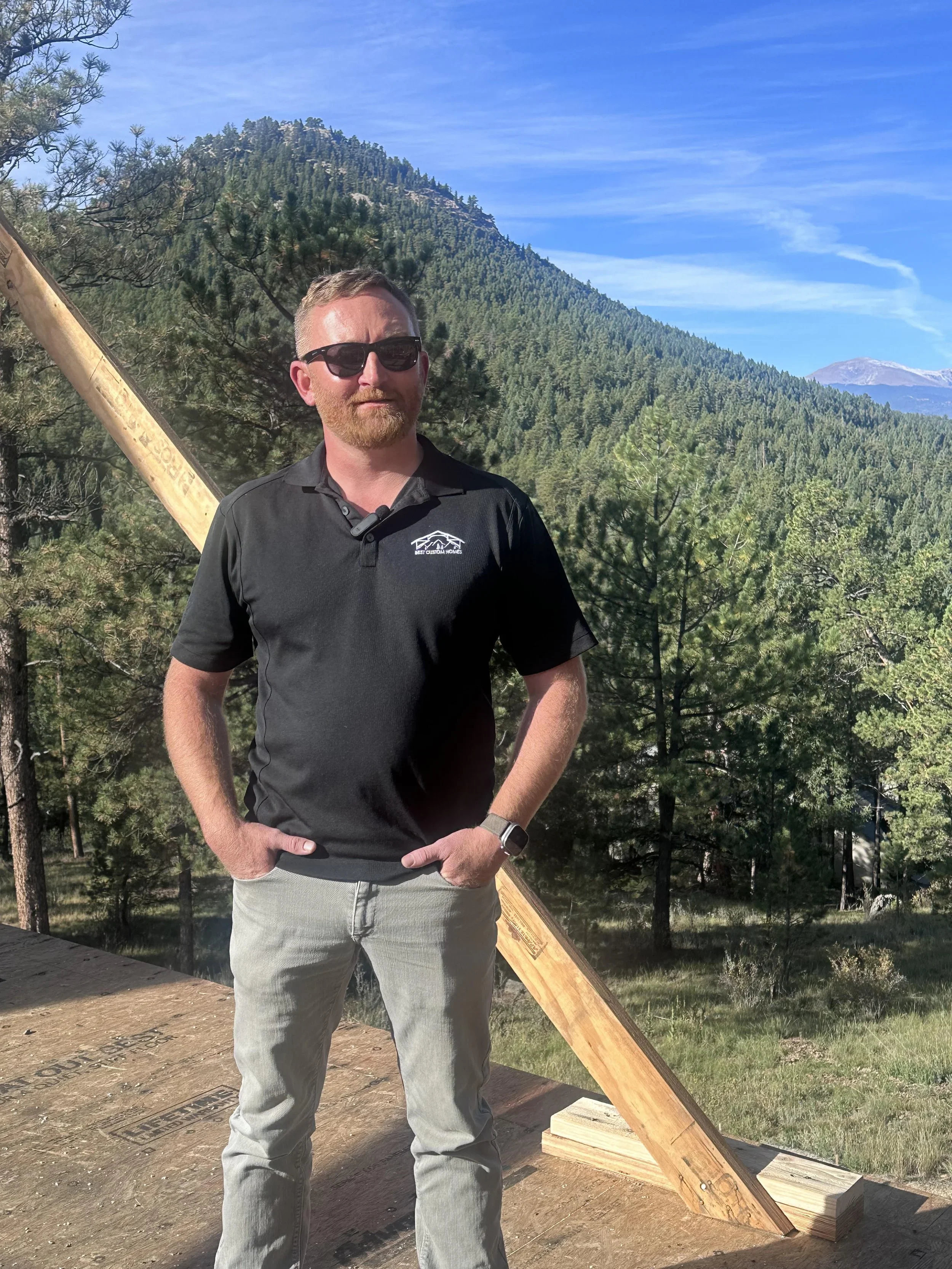 A man wearing sunglasses, a black polo shirt, and gray pants standing outdoors on a construction site, with a partially built wooden structure and a scenic mountainous forested background under a blue sky.