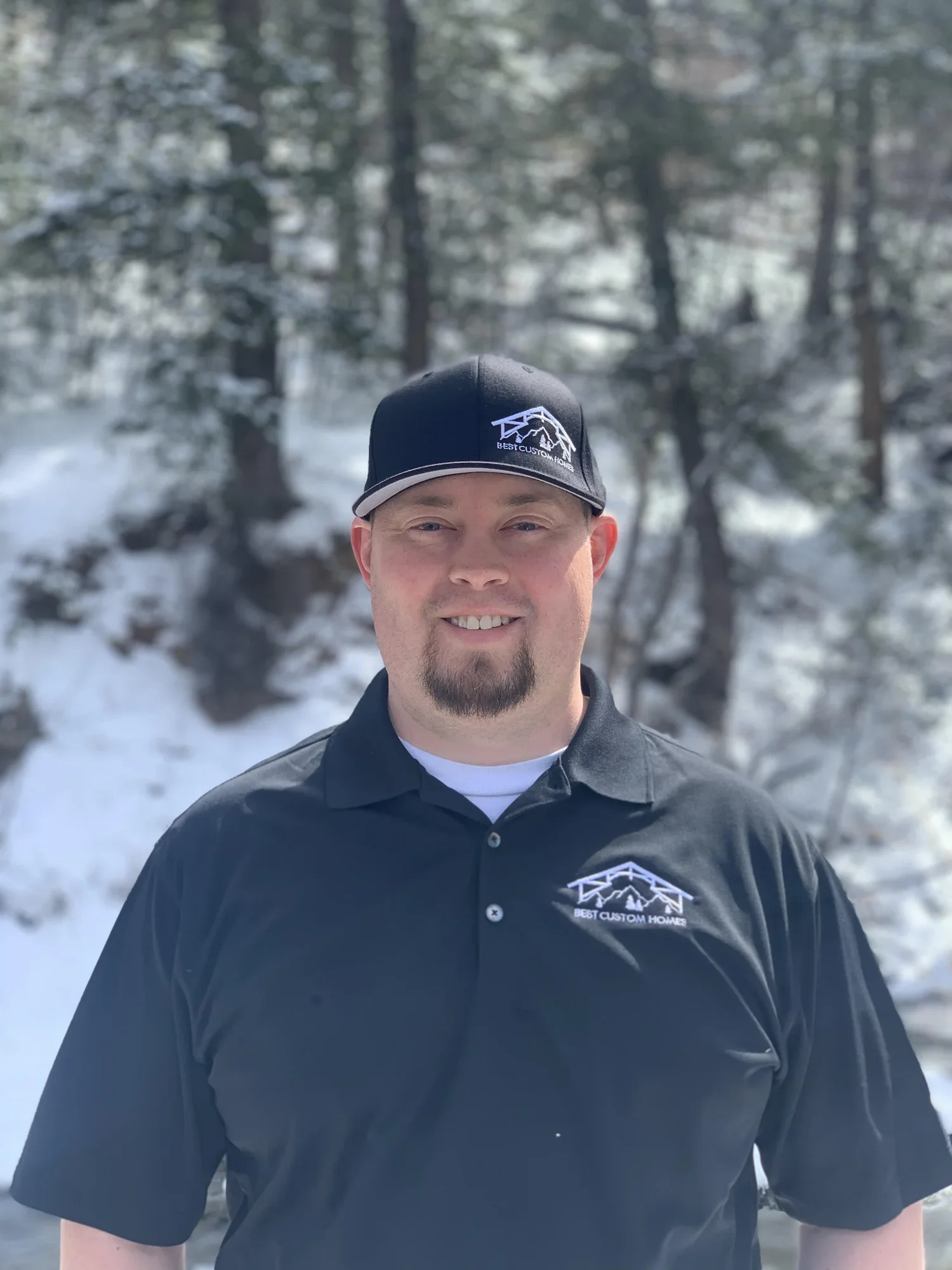 A man wearing a black baseball cap and a black polo shirt with a logo, standing outdoors in snowy terrain with trees in the background.