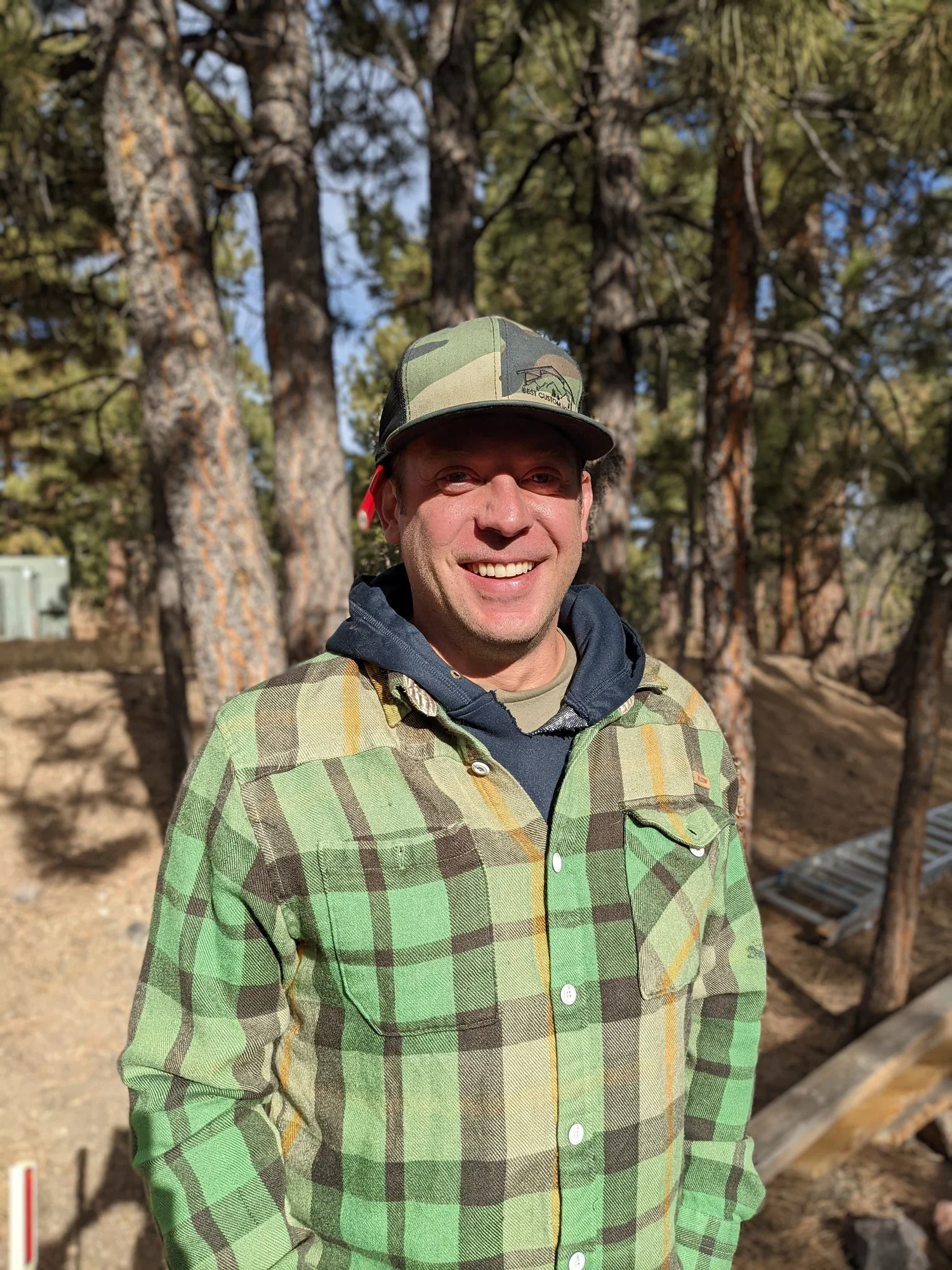 A smiling man wearing a camo cap, a plaid green and yellow shirt, and a gray hoodie, standing outdoors with tall pine trees in the background.