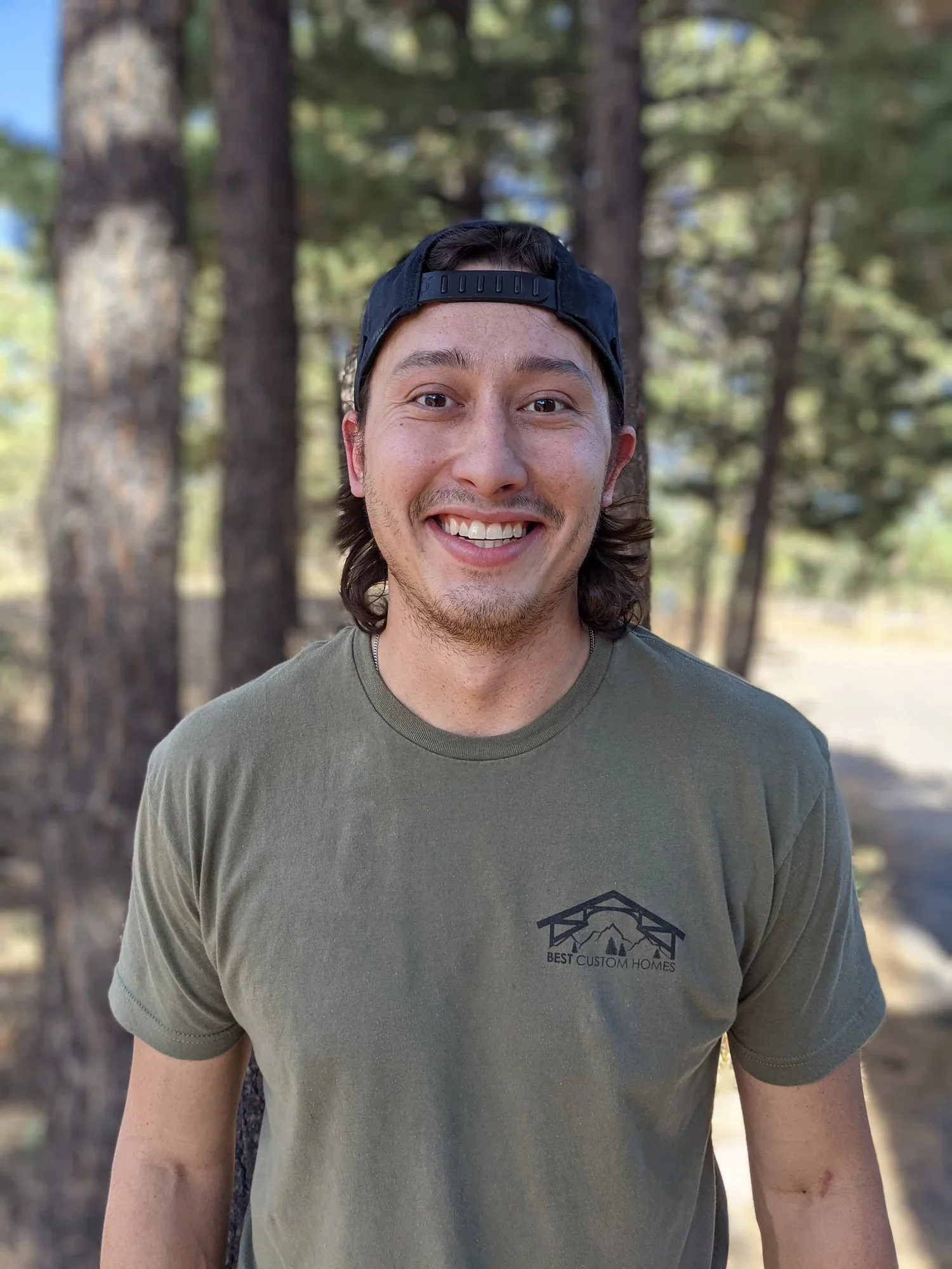 A young man smiling outdoors in a wooded area, wearing a dark baseball cap backwards and a green T-shirt with a logo that reads "Best Custom Homes."