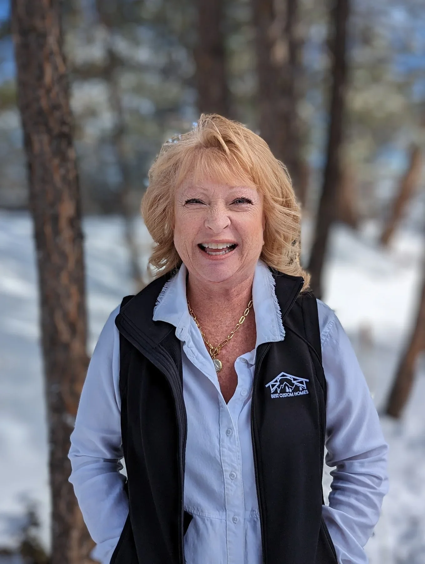 A cheerful woman with blonde hair smiling outdoors in a snowy forest, wearing a white shirt and black vest with a logo.