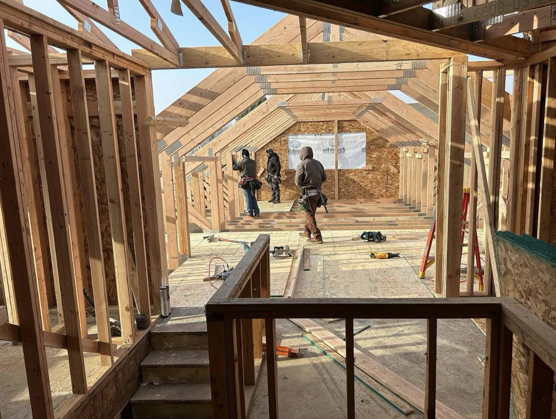 Construction workers building the wooden frame of a house interior, with unfinished stairs and beams, during daytime.