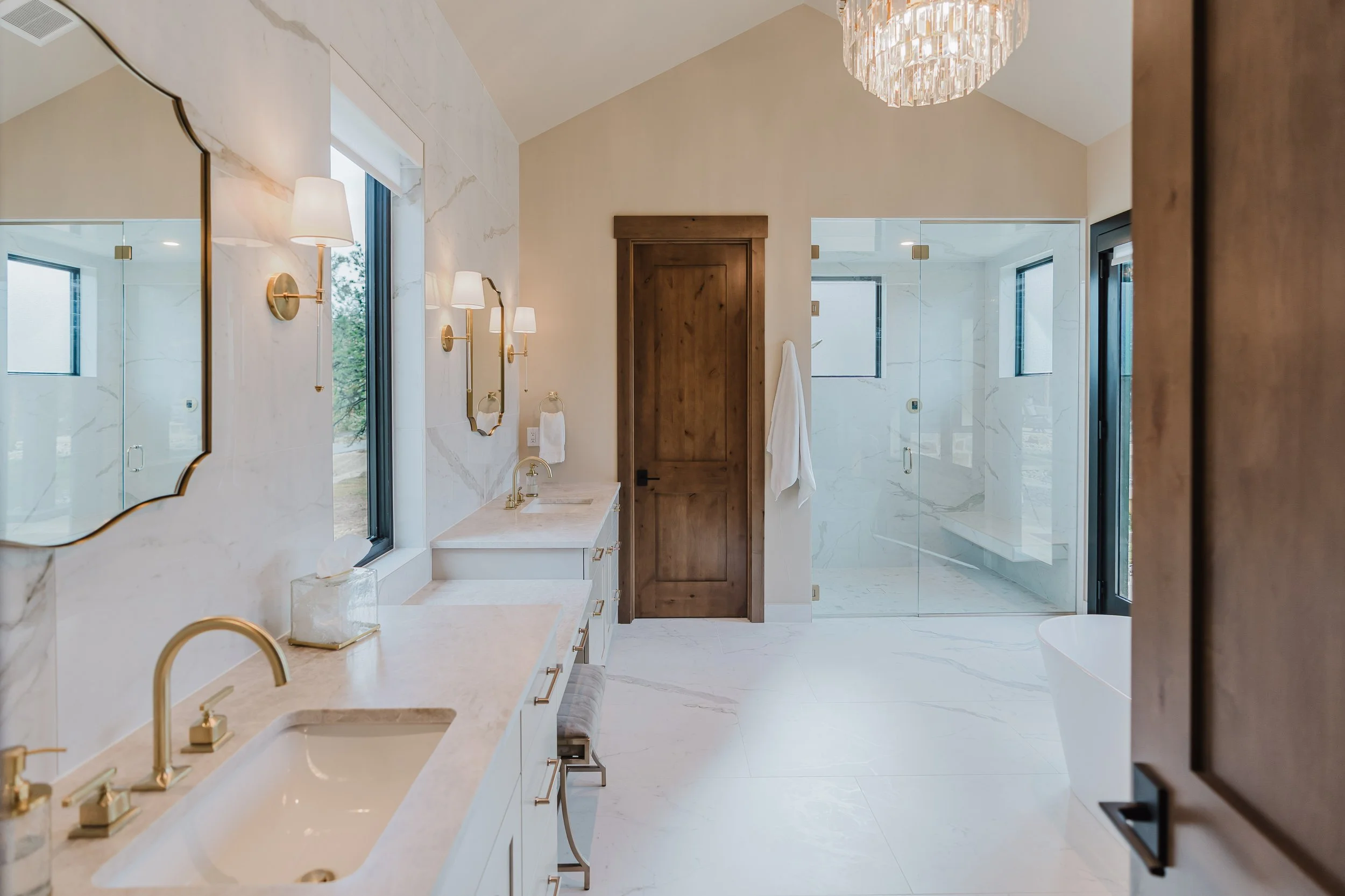 Modern bathroom featuring a double vanity with gold fixtures, wall-mounted lights, large mirror, marble countertops, wood door, and a walk-in shower with glass enclosure and marble walls.