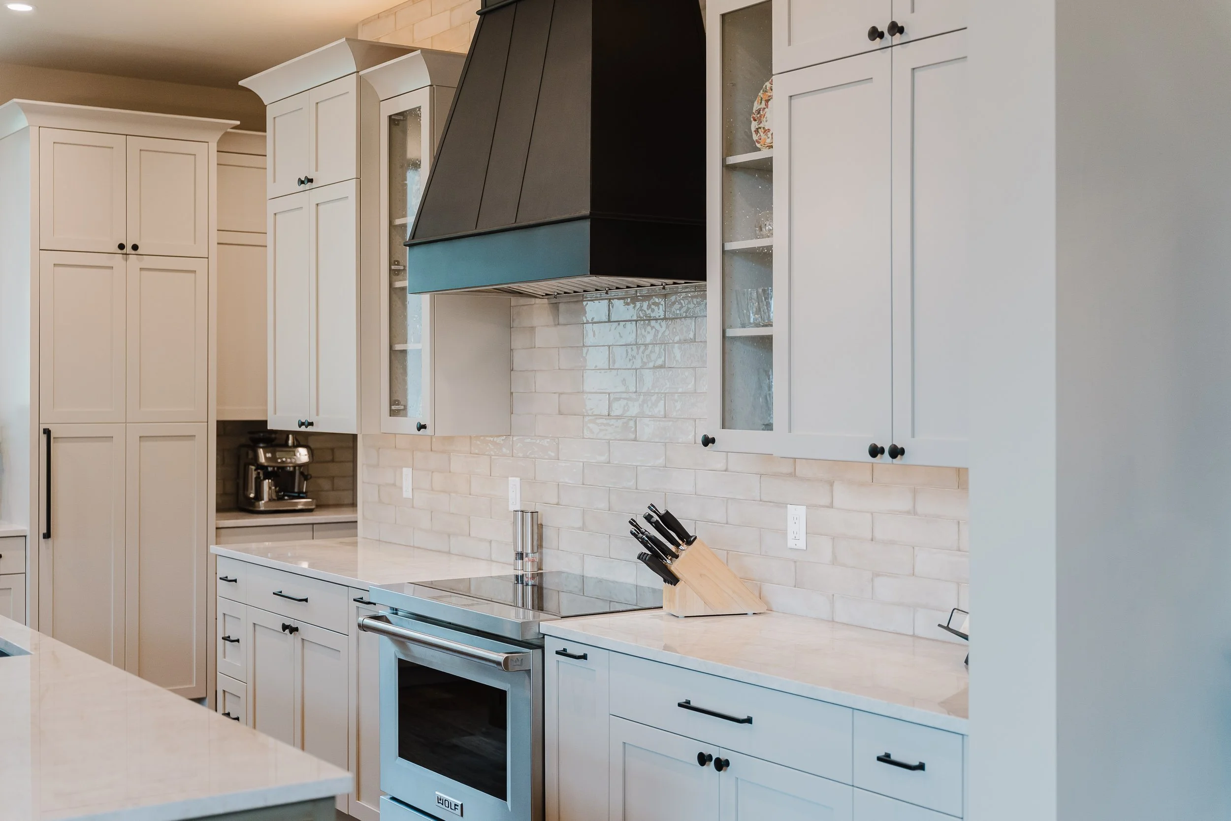 Modern kitchen with white cabinets, a stainless steel oven, a black range hood, a beige subway tile backsplash, and a knife block on the counter.