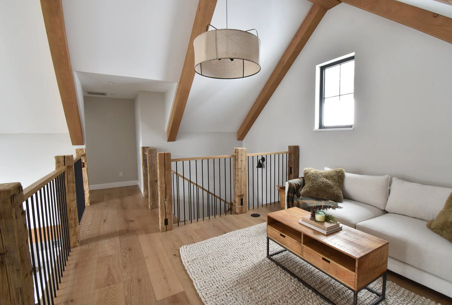 Loft interior with a white sectional sofa, wooden coffee table, cozy pillows, wood and metal railing, sloped ceiling with exposed wooden beams, window, and a large hanging light fixture.