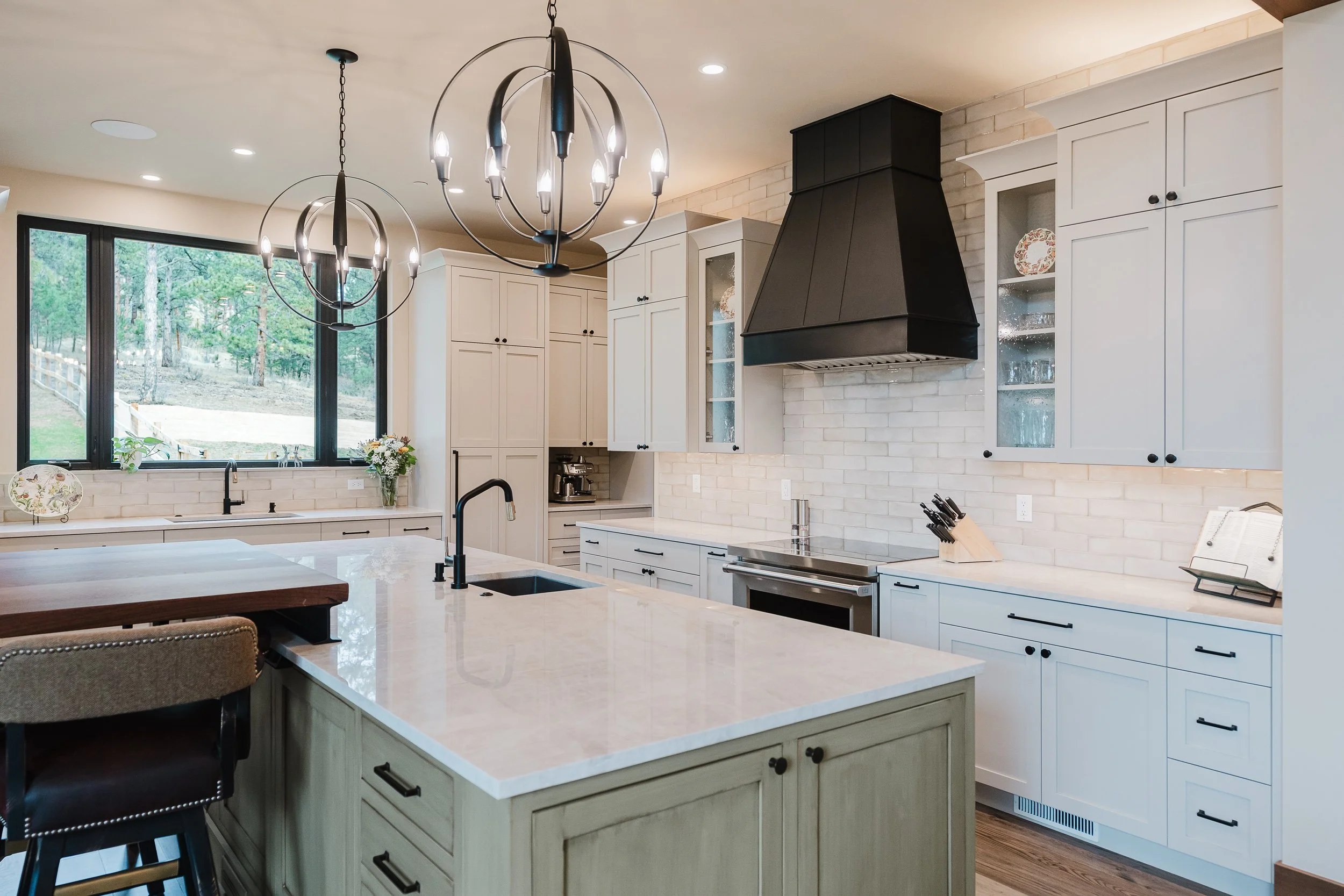 Modern kitchen with white cabinets, black hardware, black range hood, marble countertops, large window, sunlit, with a chandelier hanging above island.
